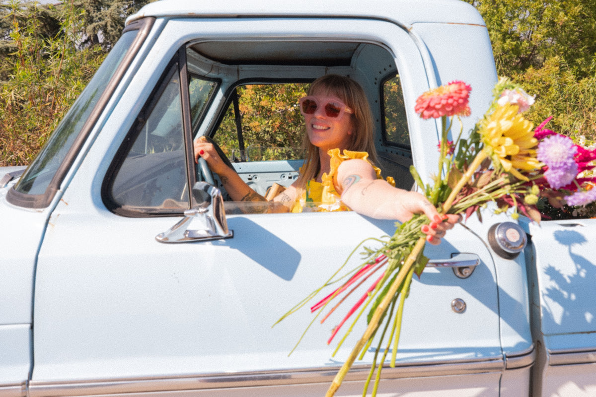 Emily smiling from the driver's seat of the baby-blue flower truck, holding a bright bouquet out the window